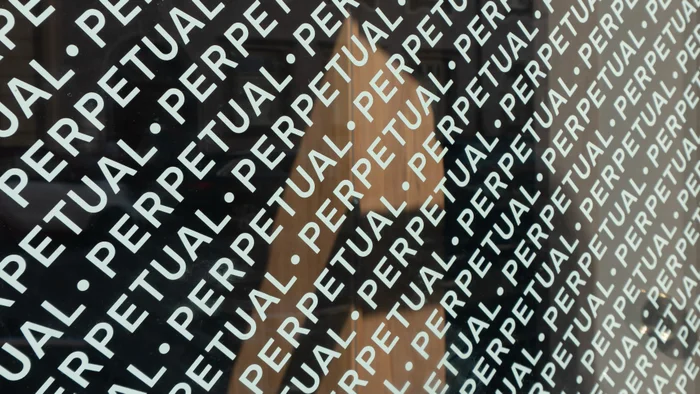 Close-up angled shot of dark glass surface featuring repeated white typographic text reading PERPETUAL with dot separators