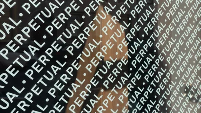 Close-up angled shot of dark glass surface featuring repeated white typographic text reading PERPETUAL with dot separators