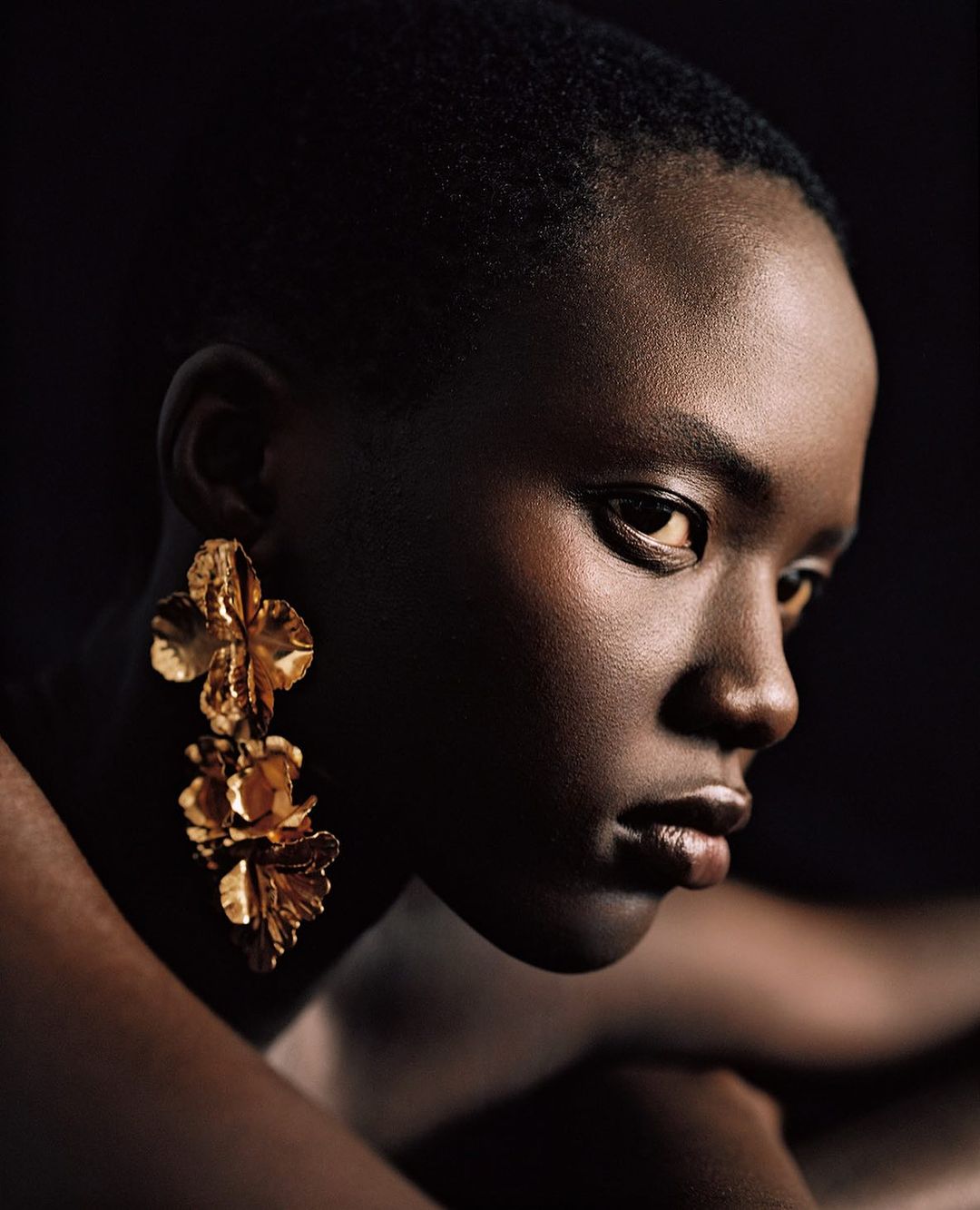 Close-up studio portrait of an adult Black woman with short hair, wearing large gold floral earrings, against a dark backdrop.