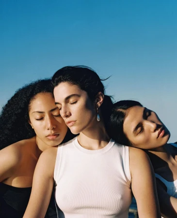 Three women posing closely together with closed eyes against a clear blue sky background outdoors