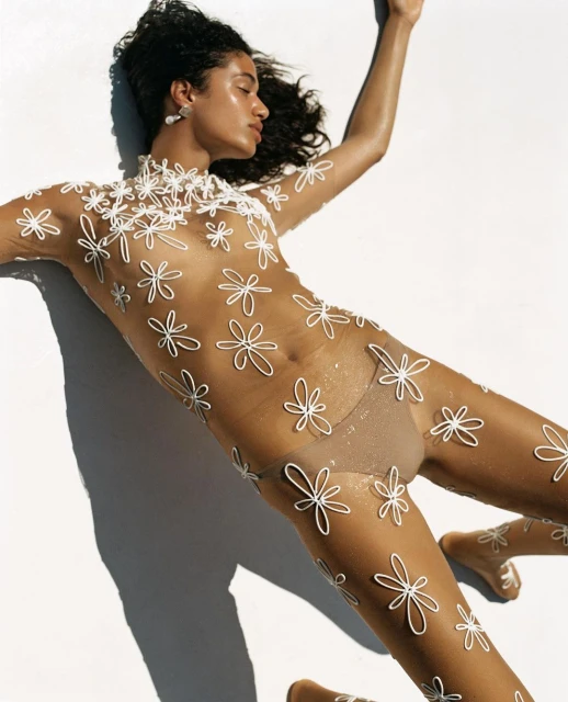 Woman lies on a white surface wearing a sheer body suit with 3D flower embellishments. Studio lighting illuminates the scene.