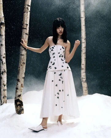 An Asian woman in a white corset dress with black insect patterns stands in a snowy birch tree forest setting for a fashion editorial.