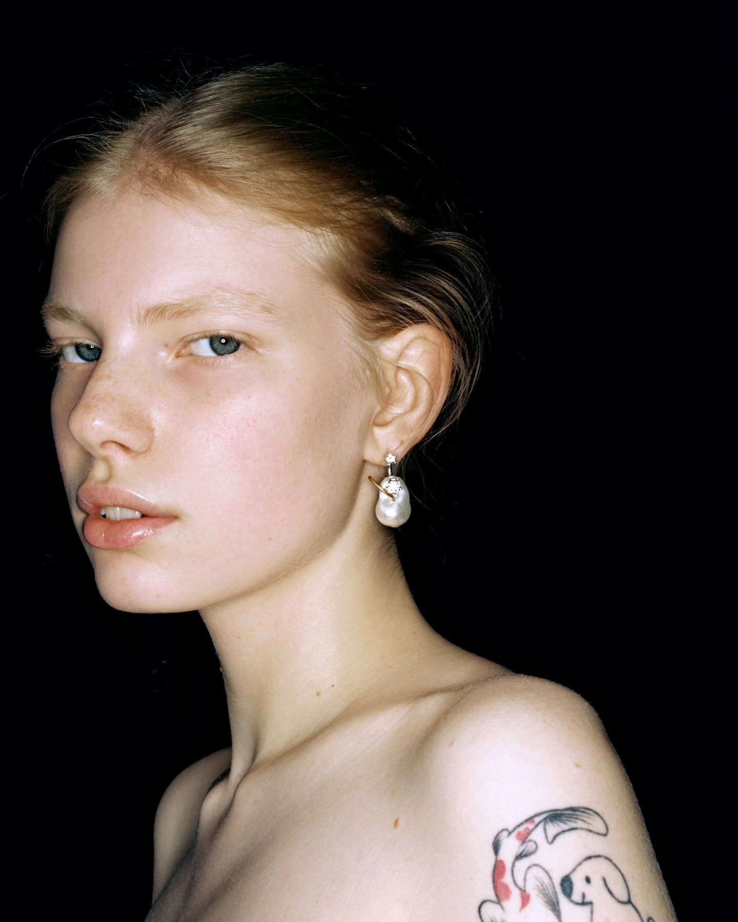 Close-up studio portrait of a fair-skinned young woman with light hair, wearing a pearl earring, against a black background.