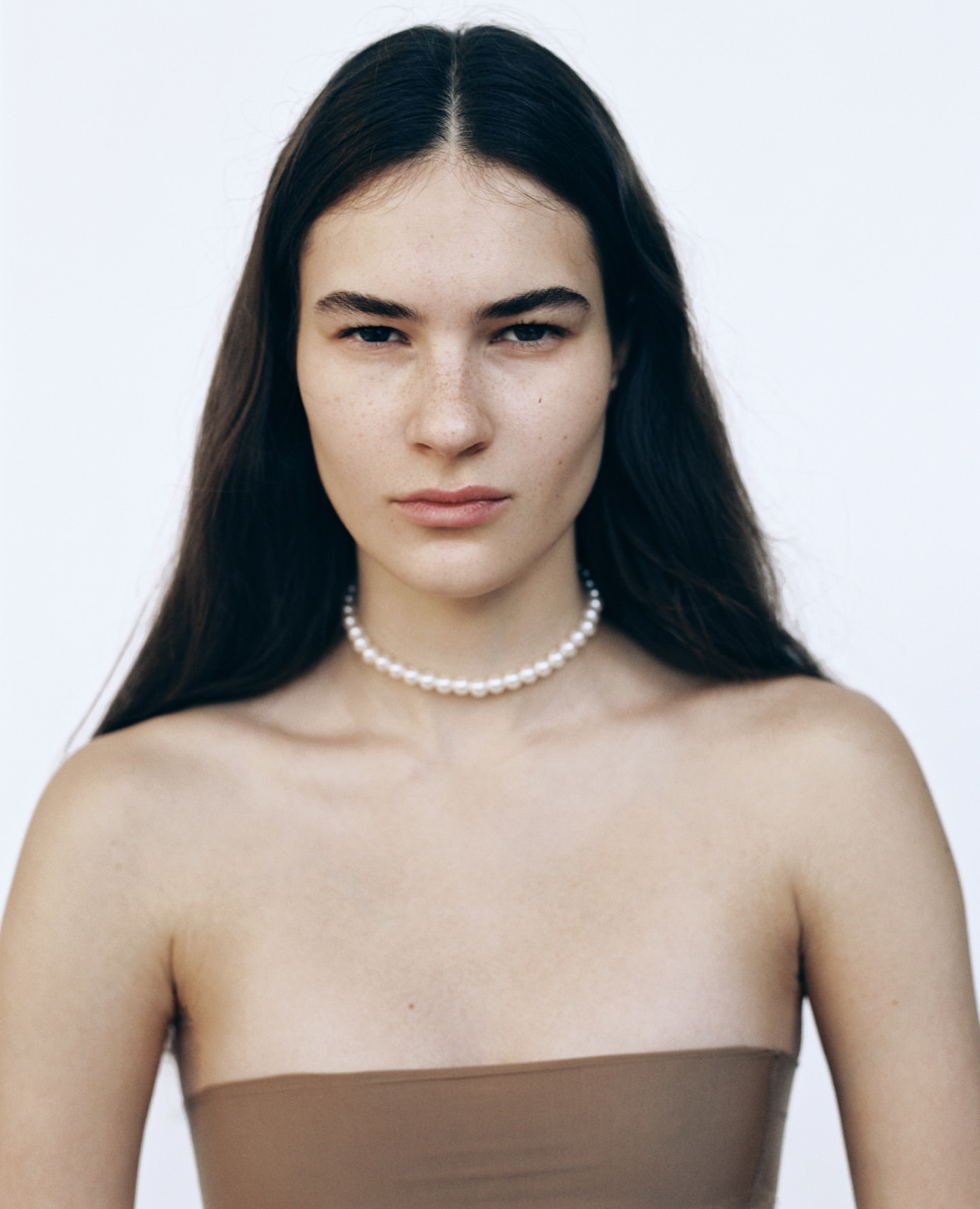 Close-up portrait of a young woman with long dark hair wearing a pearl choker against a plain white backdrop in soft, diffused lighting.
