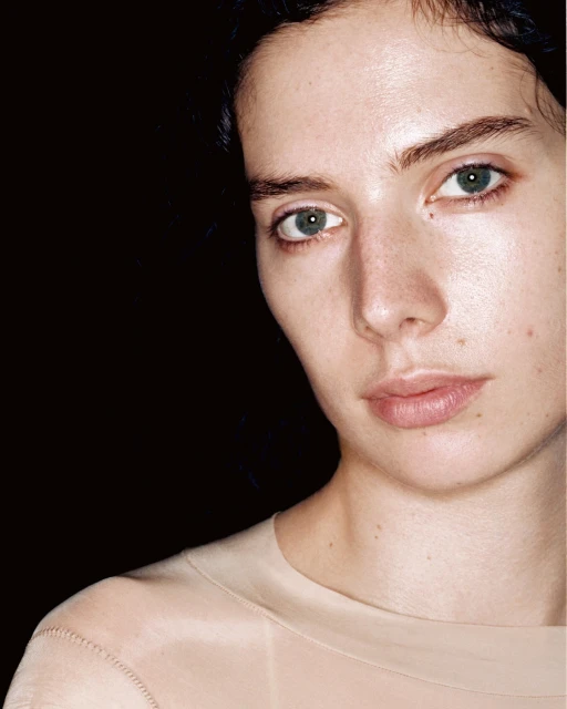 Close-up portrait of a young adult woman with fair skin and green eyes in a peach-colored shirt against a dark background.