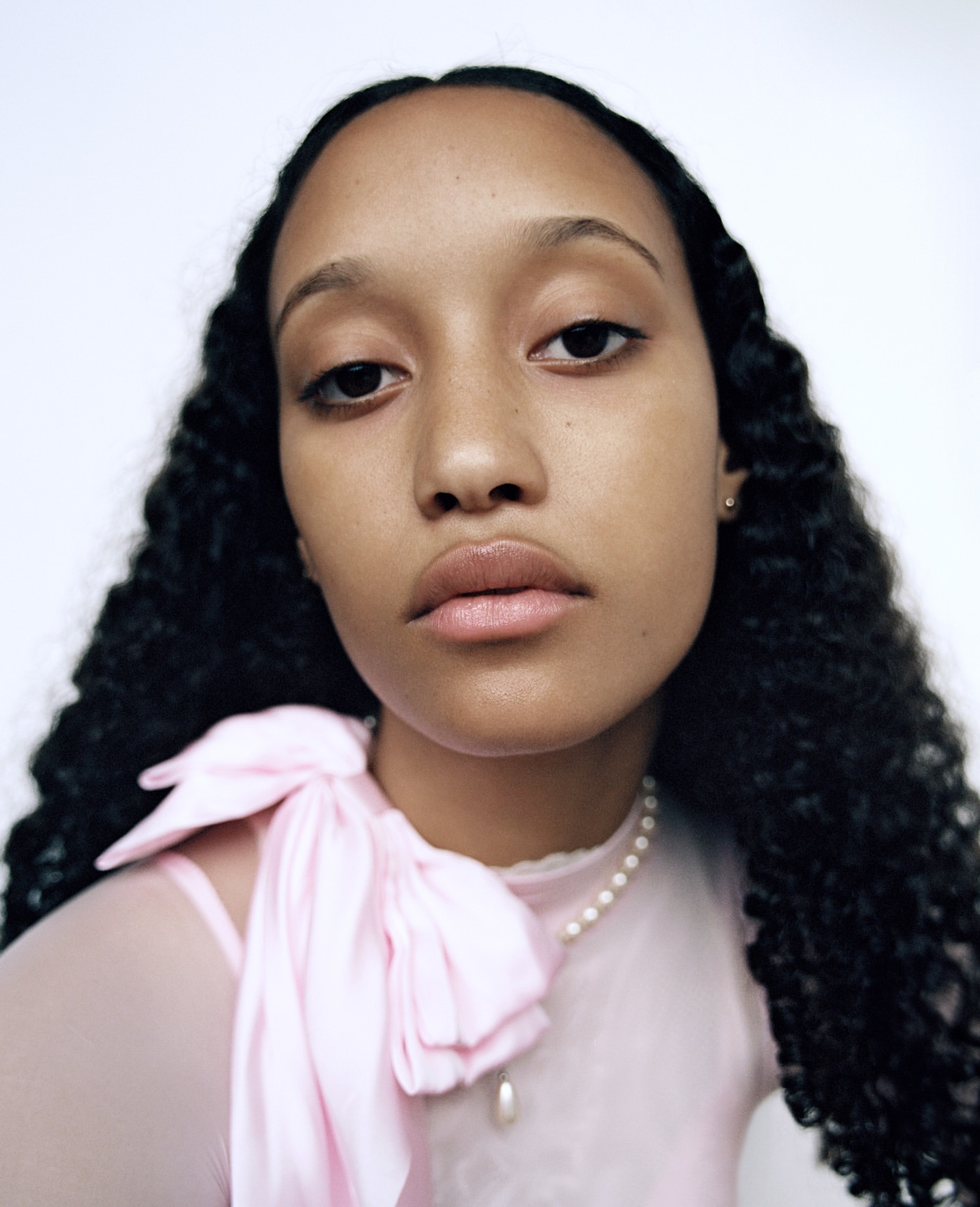 Close-up portrait of a young adult female with dark curly hair, wearing a sheer pink top with a bow and a pearl necklace against a white background.