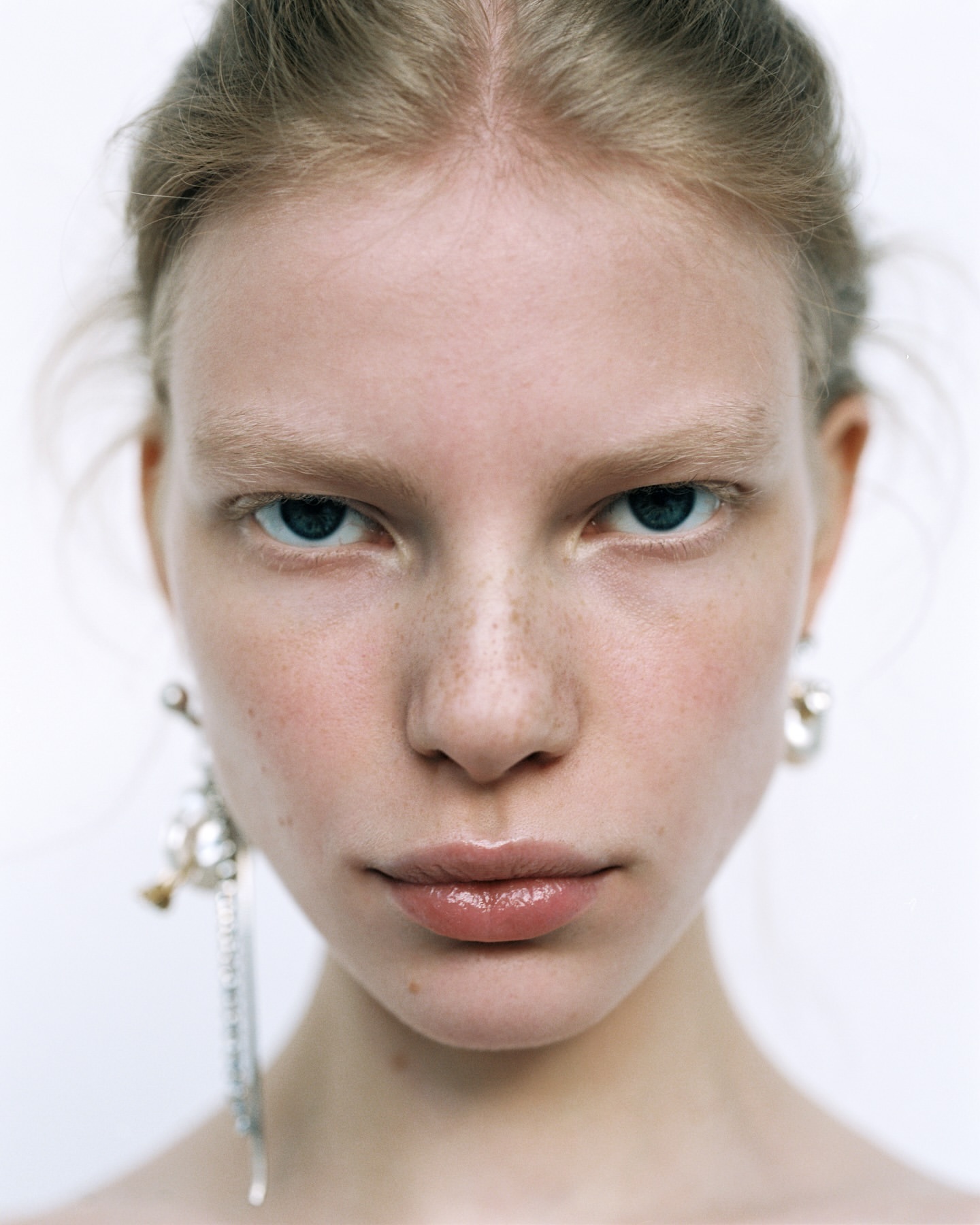 Close-up portrait of a fair-skinned woman with blue eyes, freckles, and light blonde hair, wearing dangle earrings against a white backdrop.