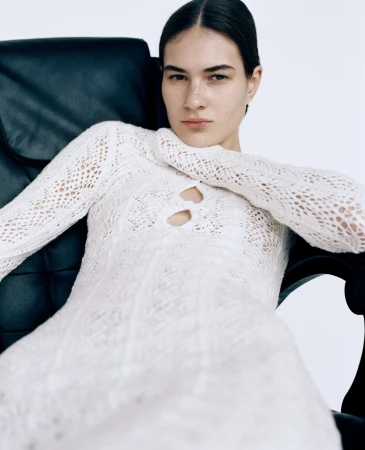 Model in white knitted dress seated in a black leather armchair against a plain white background