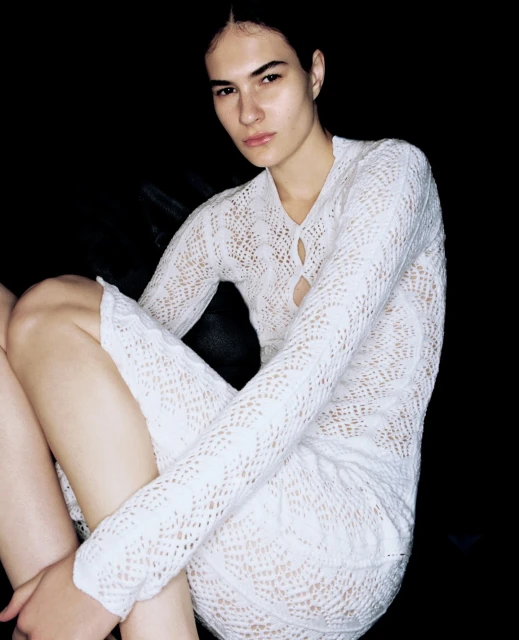 Portrait of a young adult woman with dark hair wearing a white crocheted dress, sitting indoors against a black background in soft lighting.