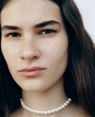 Close up portrait of a young woman with dark hair and freckles wearing a pearl necklace against a plain white background