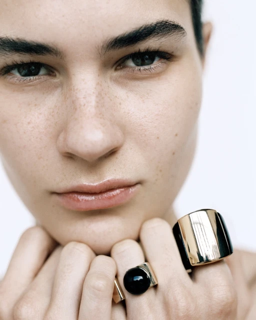 Close-up portrait of a woman with fair skin and freckles wearing chunky gold rings against a plain white background in a beauty shot.