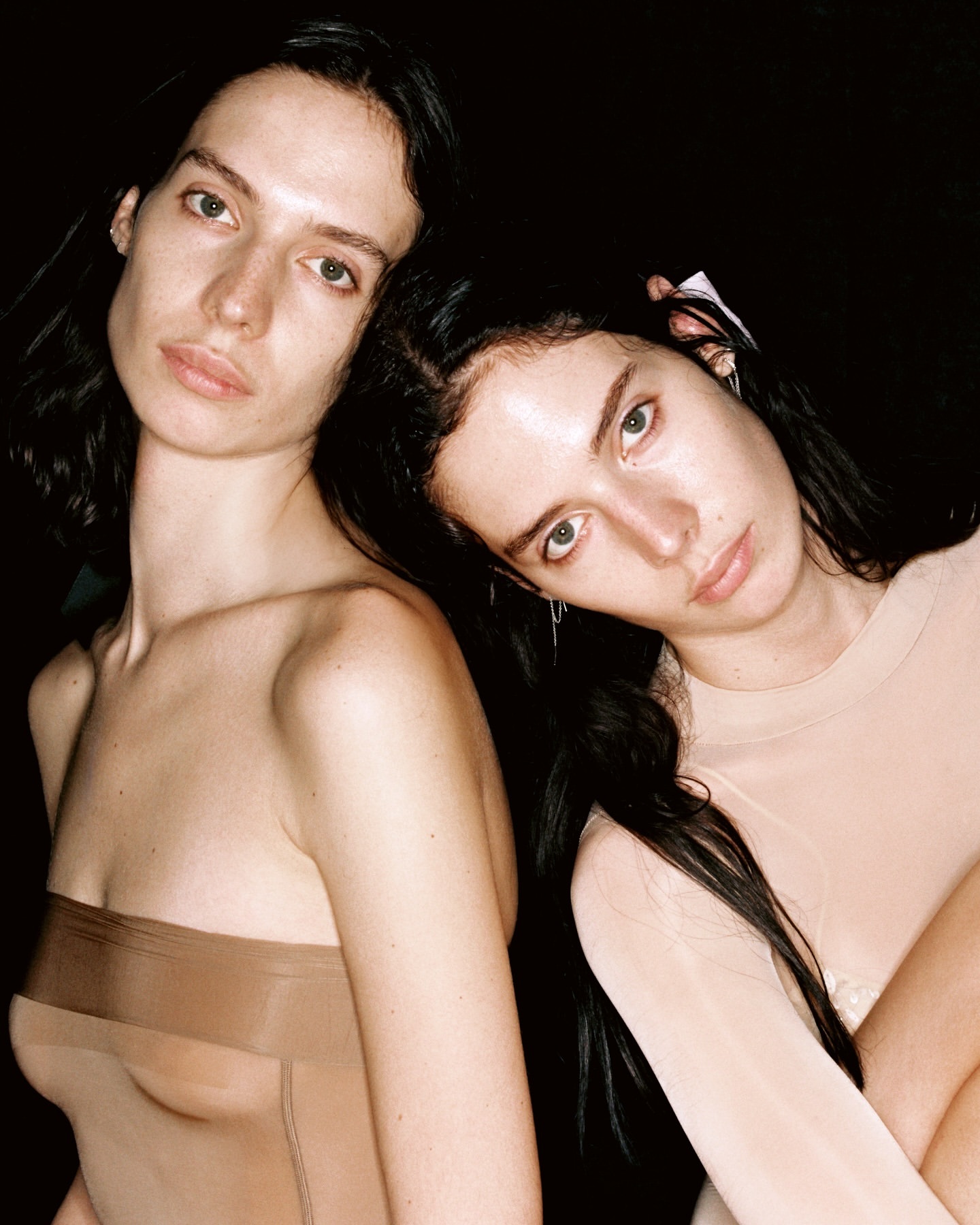 Two women in neutral tones, close-up portrait. One woman is fair-skinned and in a beige top, the other is partially undressed in a brown tone.