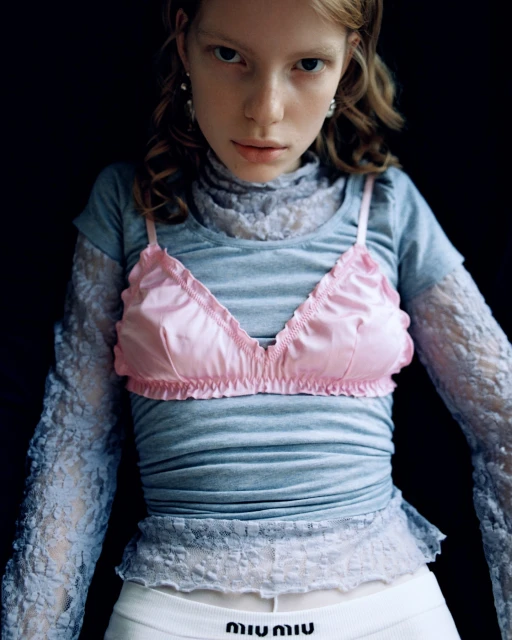 Portrait of a fair-skinned young woman with blonde hair in a layered outfit with a pink bra and white bottoms with logo.