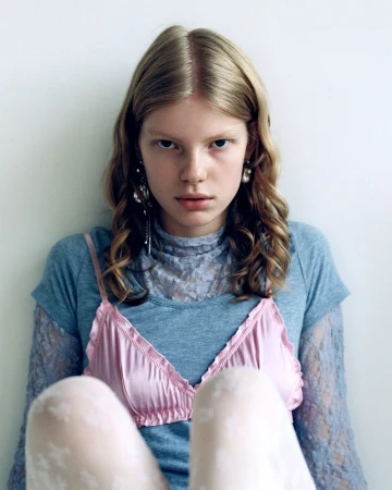 Close-up portrait of a young blonde woman in a pink slip top and blue lace top against a plain wall.