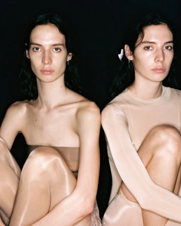 Two women sit closely in front of a black backdrop. One has a ribbon in her hair and wears a tan long-sleeved top.