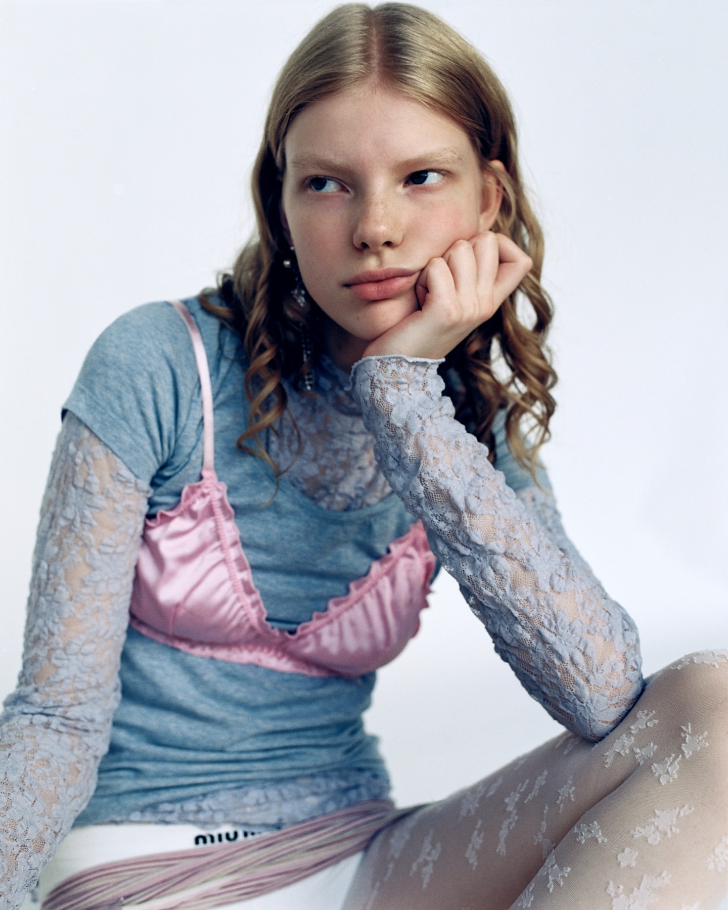 Young woman sits in a studio wearing a blue shirt, satin bra, and patterned leggings, with a hand on her cheek looking sideways.