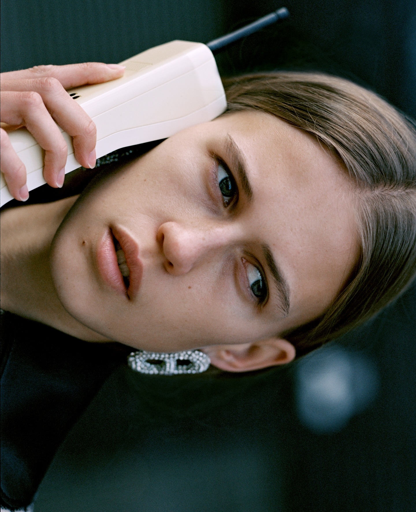 Close-up studio portrait of a young woman using a large vintage cell phone, adorned with sparkling earrings and dark clothing.