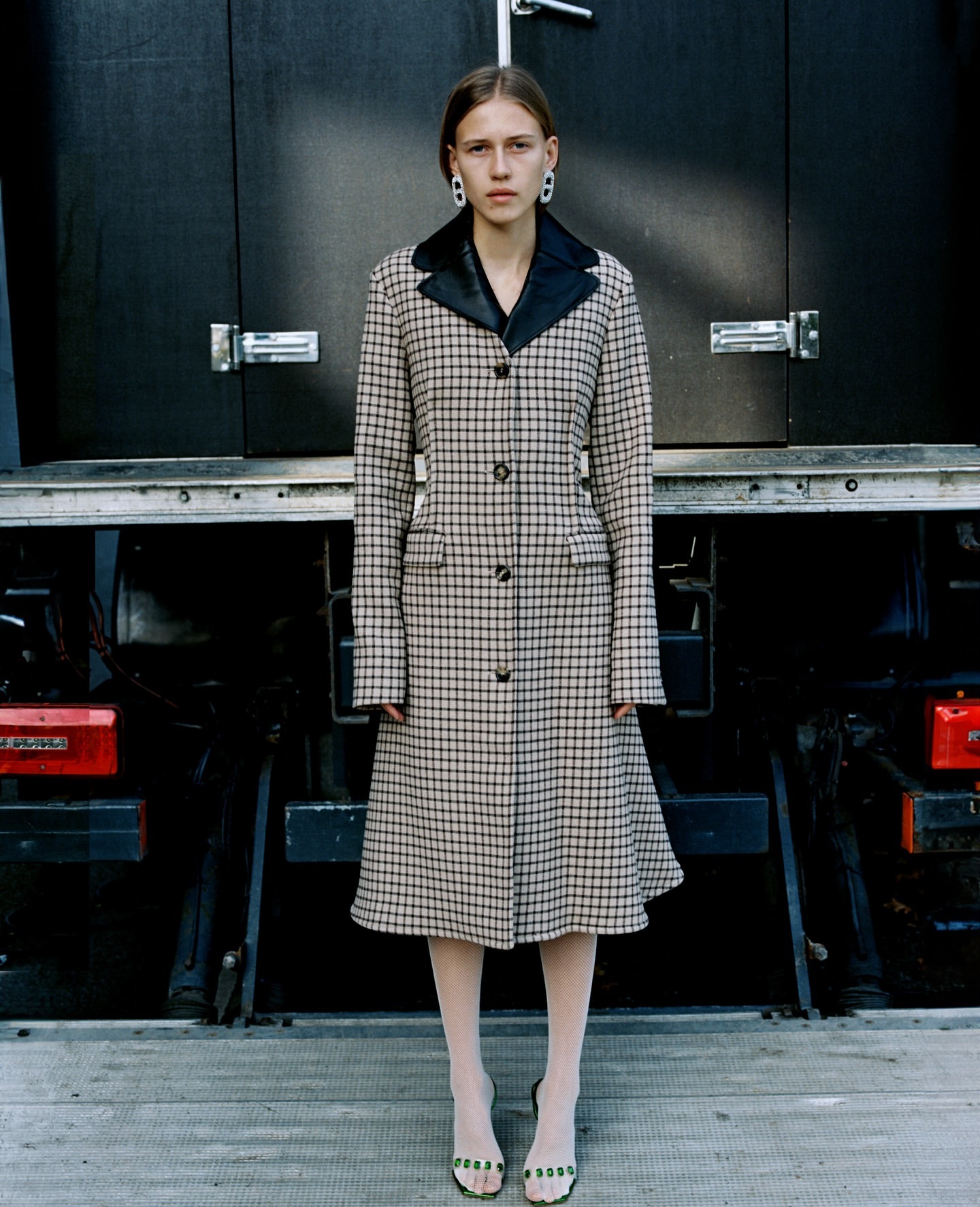 A young woman poses in a checkered coat and light tights against a dark truck backdrop, showcasing fashion and urban style.