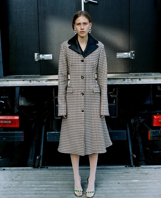 A young woman poses in a checkered coat and light tights against a dark truck backdrop, showcasing fashion and urban style.