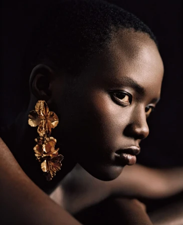 Close-up portrait of a Black woman wearing large textured gold floral drop earrings against a dark background.