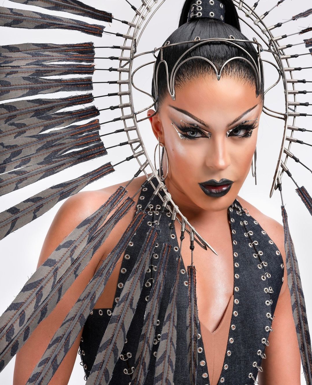 Close-up of a striking drag queen with elaborate eye makeup, dark lipstick, and a unique feathered headpiece on a plain white background.