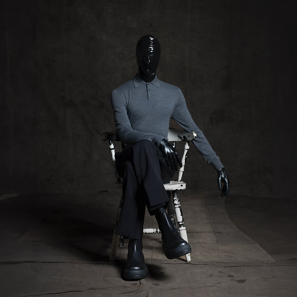 Mannequin in gray sweater, black pants and boots sitting on a white chair against dark gray backdrop in an indoor setting.