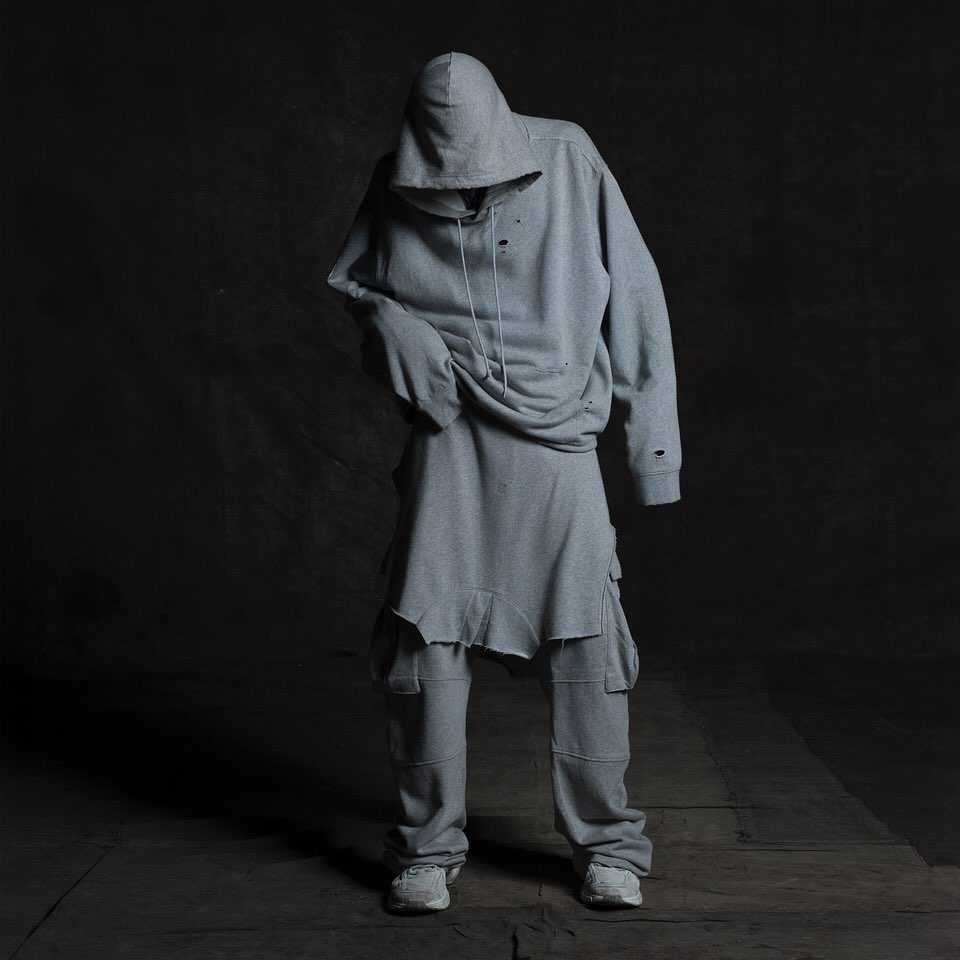 Person standing in grey ripped hoodie and pants, lit with spotlight, studio setting. Fashion shot for editorial or clothing design.