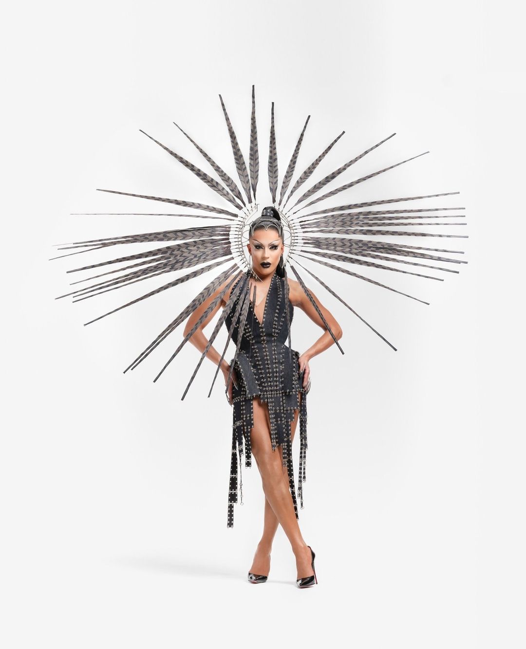 Drag queen posing in a studded black dress with a sunburst headpiece against a white backdrop, captured in a full shot.