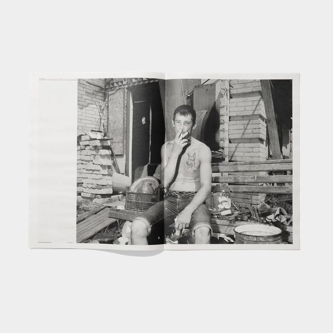 Black and white photo of shirtless man smoking cigarette in dilapidated urban setting with brick walls and debris in open book.