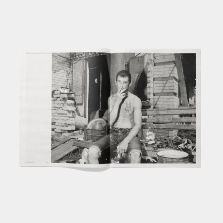 Black and white photo of shirtless man smoking cigarette in dilapidated urban setting with brick walls and debris in open book.