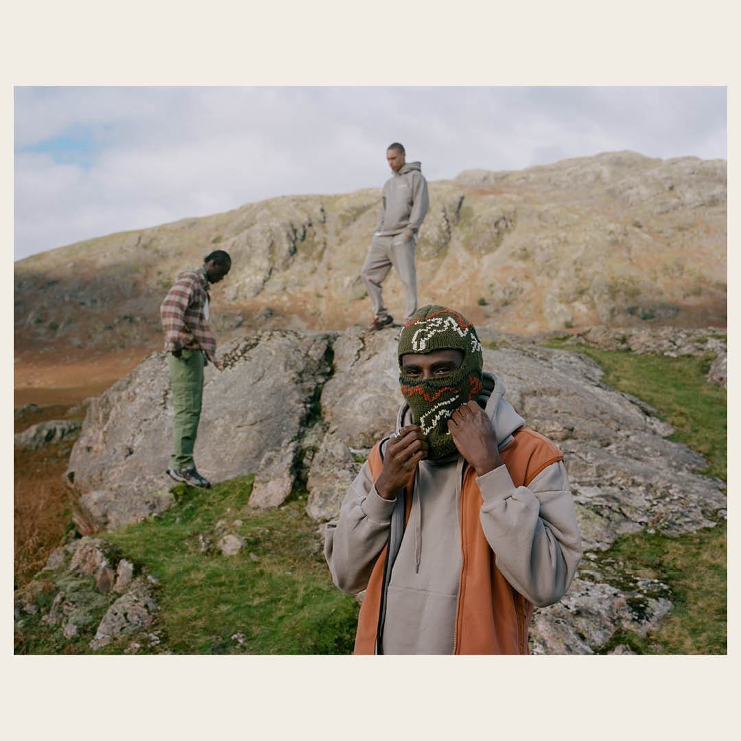 Three men pose in a mountain landscape, one wearing a patterned balaclava and an orange vest over a grey hooded sweatshirt.