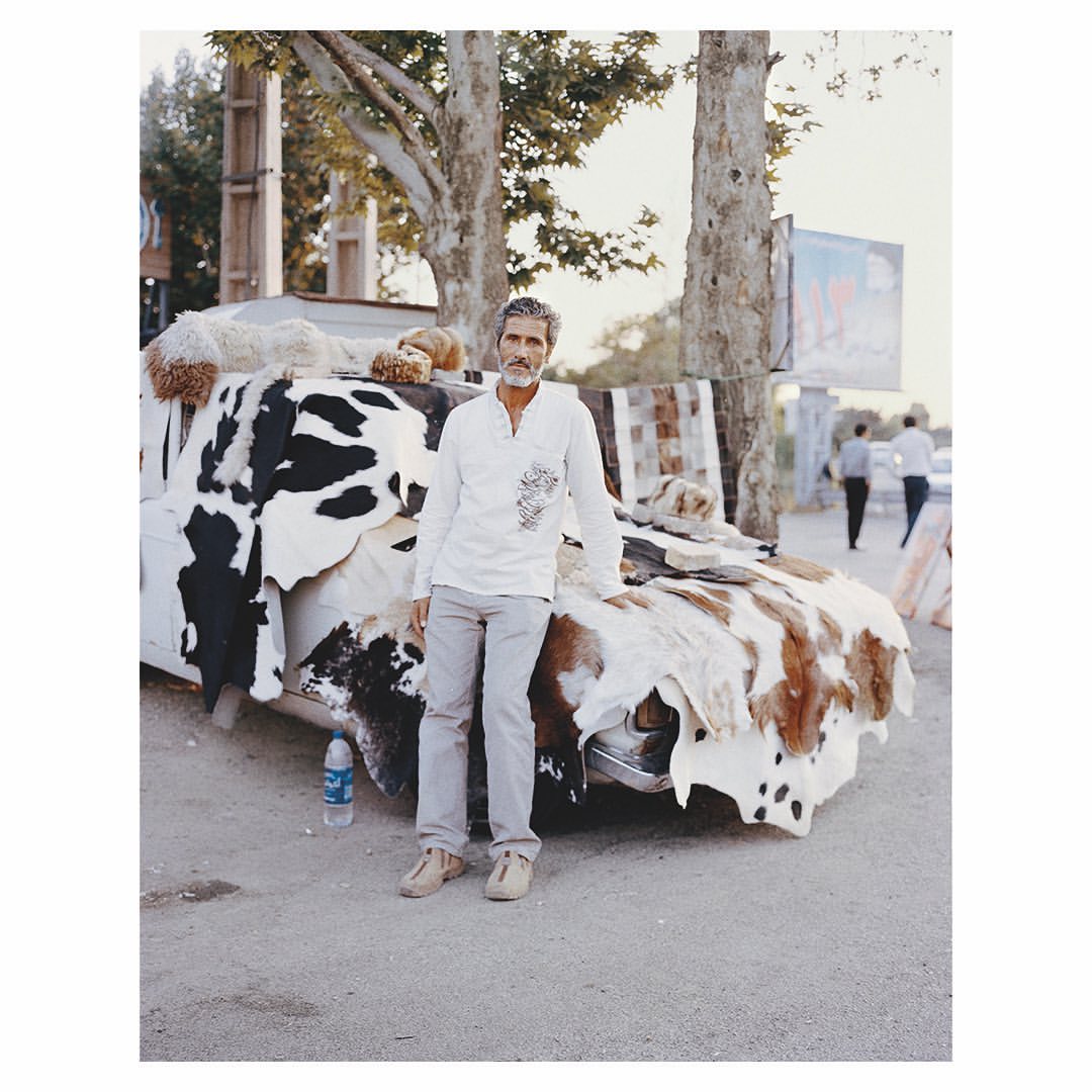 Mature man stands next to vintage car adorned with animal hides on a street with trees, a billboard, and other people in the background.