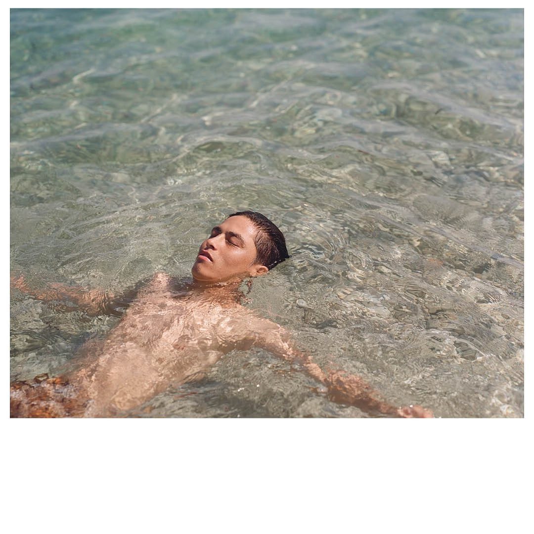 Man floating on his back in clear, shallow water, eyes closed, arms outstretched to the sides, wearing trunks, serene expression.