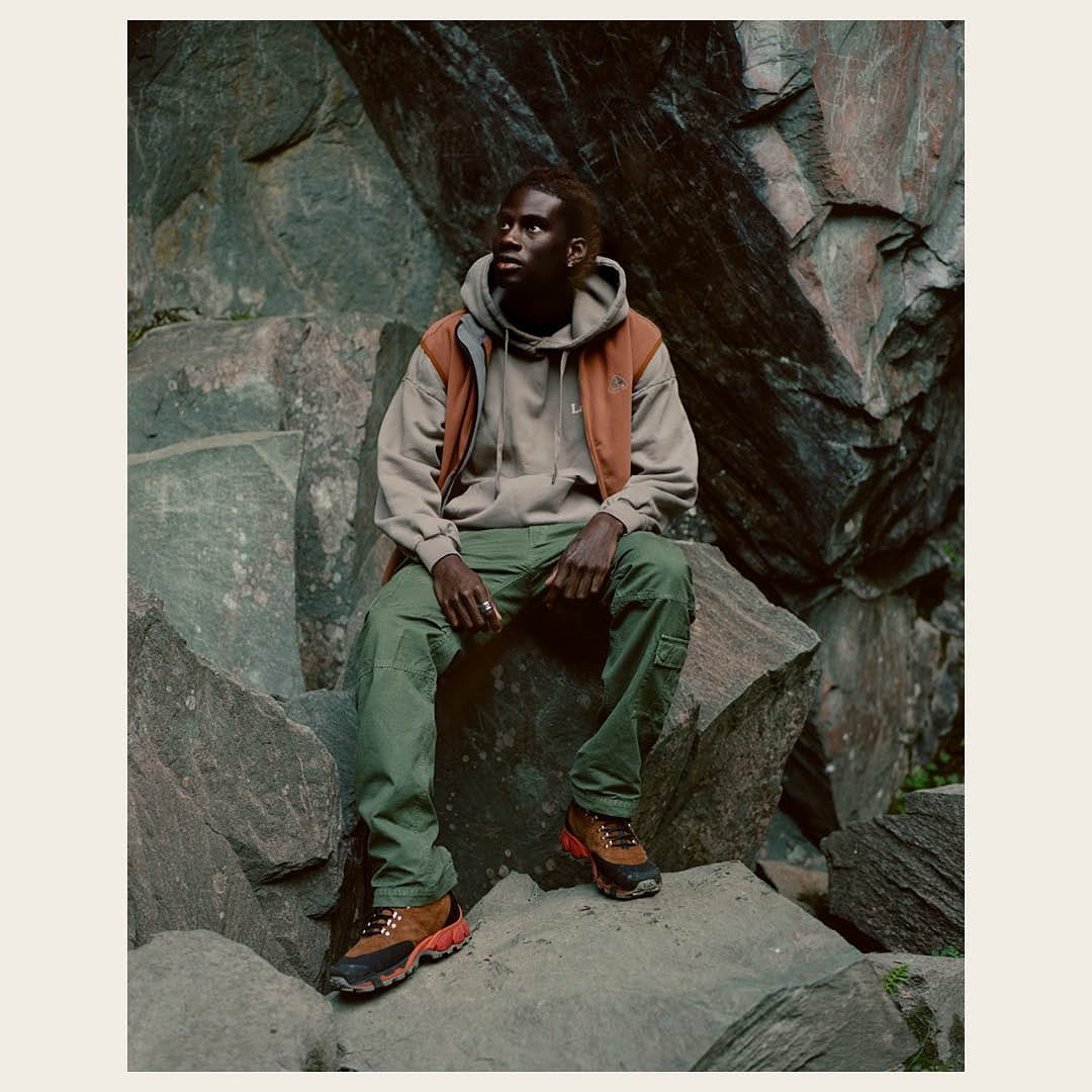 Male model in casual outerwear sits on a rock in an outdoor, natural rock setting, wearing earth-toned layers and outdoor hiking footwear.