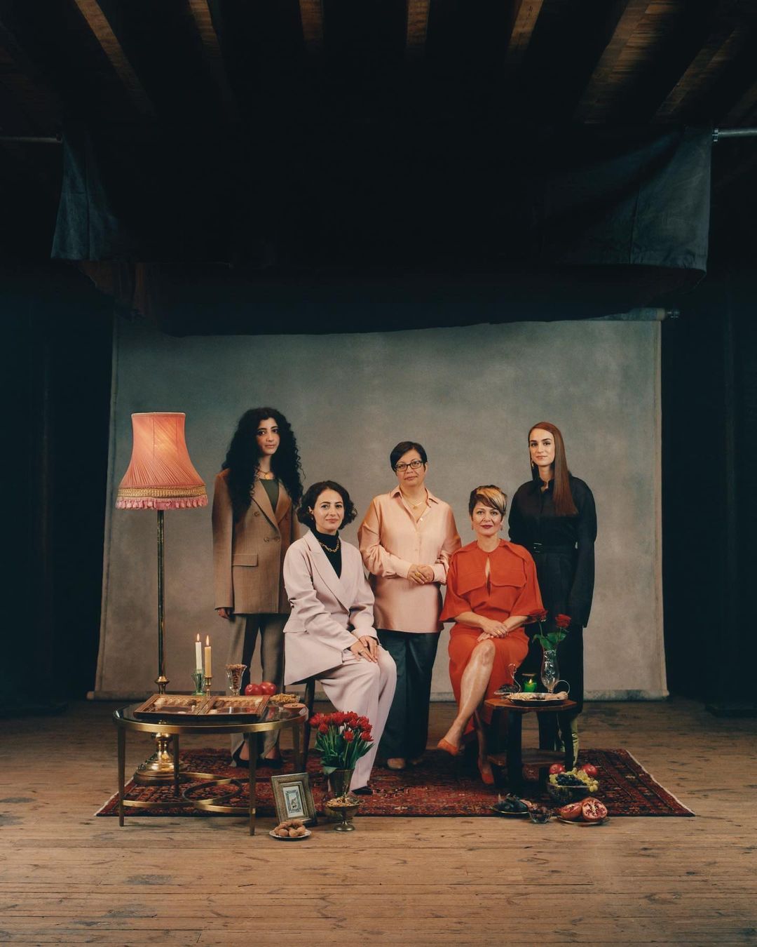 Group portrait of five adult women in various dresses and suits, posing indoors with vintage-style decor and props in soft, ambient light.