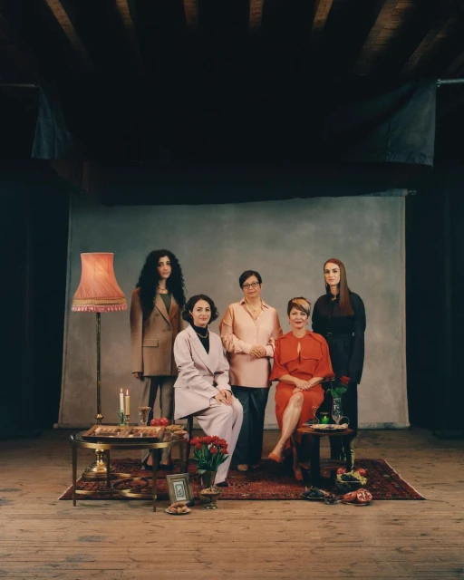 Group portrait of five adult women in various dresses and suits, posing indoors with vintage-style decor and props in soft, ambient light.