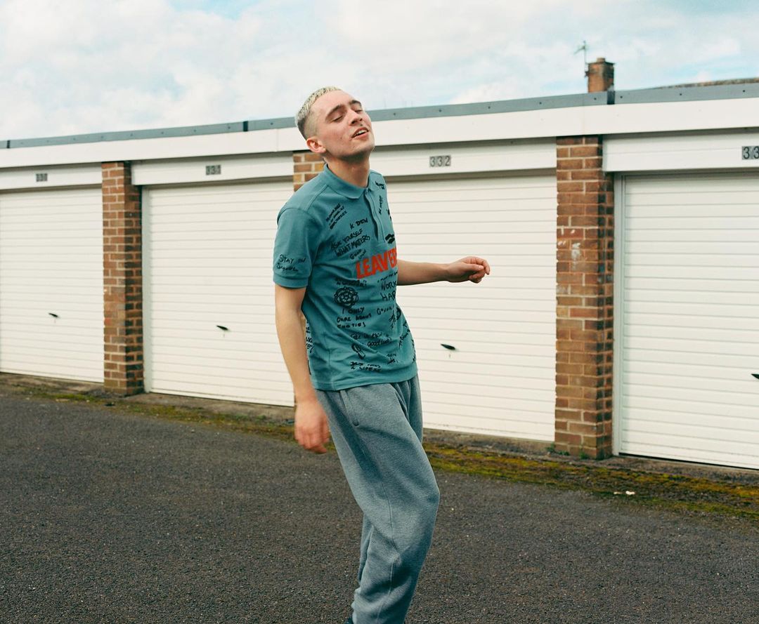 Young adult male with buzzcut in teal polo shirt and grey sweatpants stands on an asphalt surface in front of garage doors.
