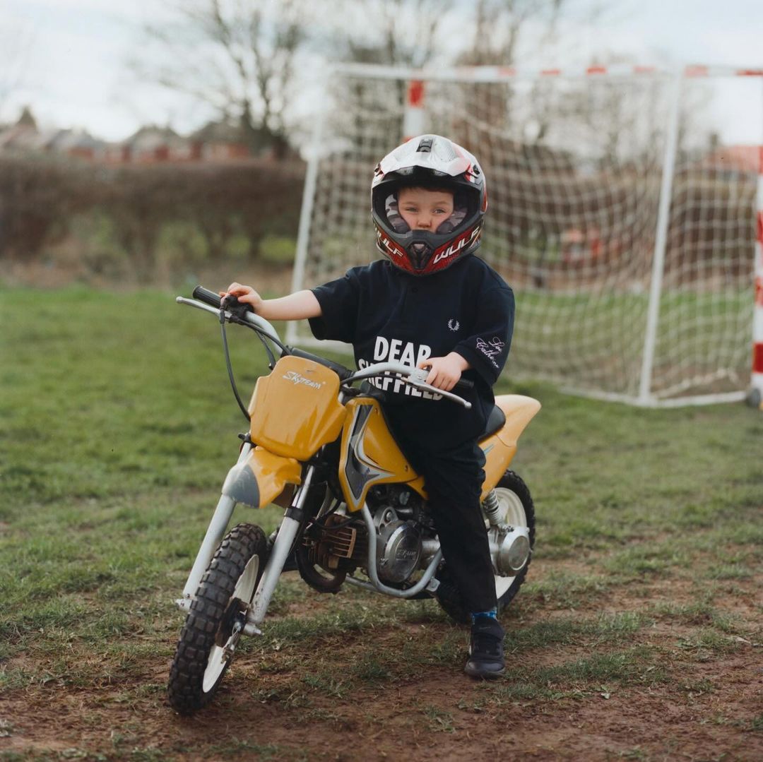 A child sits on a yellow dirt bike in a grassy yard, wearing a helmet and black clothing. A soccer goal net is visible in background.
