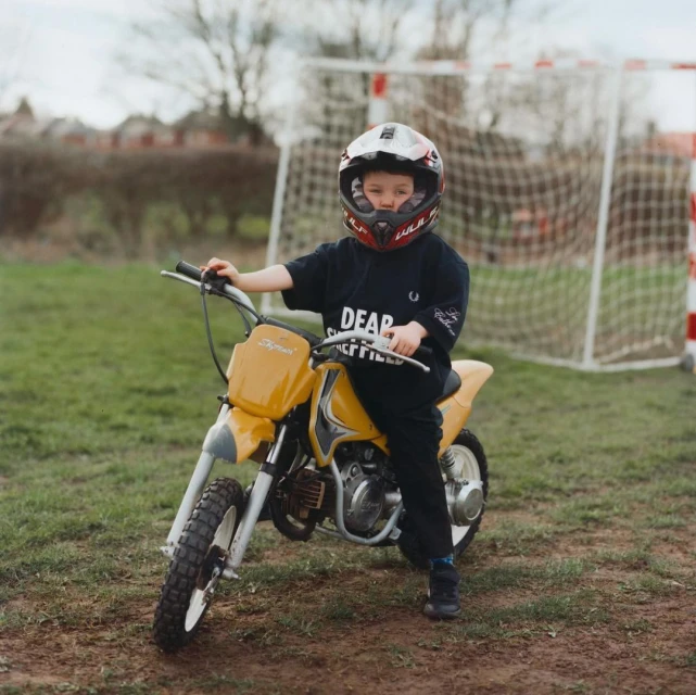 A child sits on a yellow dirt bike in a grassy yard, wearing a helmet and black clothing. A soccer goal net is visible in background.