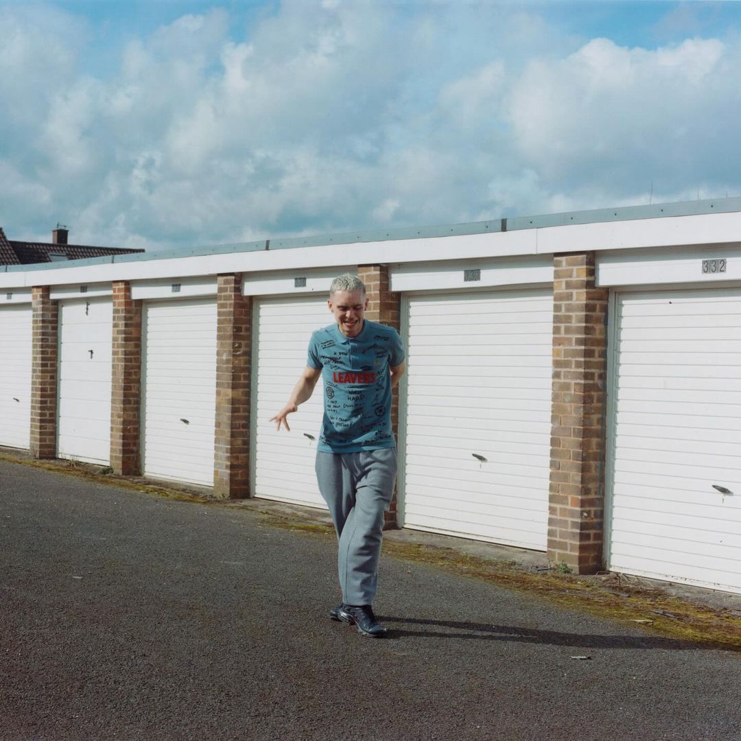 Adult male walking along a row of white garages under a cloudy sky, captured in a medium shot with natural lighting.