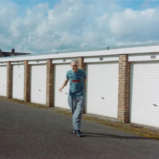 Adult male walking along a row of white garages under a cloudy sky, captured in a medium shot with natural lighting.