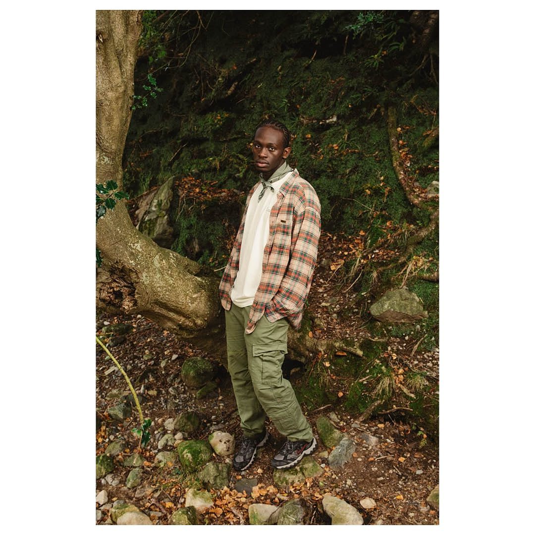 Man in plaid shirt and cargo pants poses in an outdoor setting with a mossy boulder, surrounded by lush green foliage and natural light.