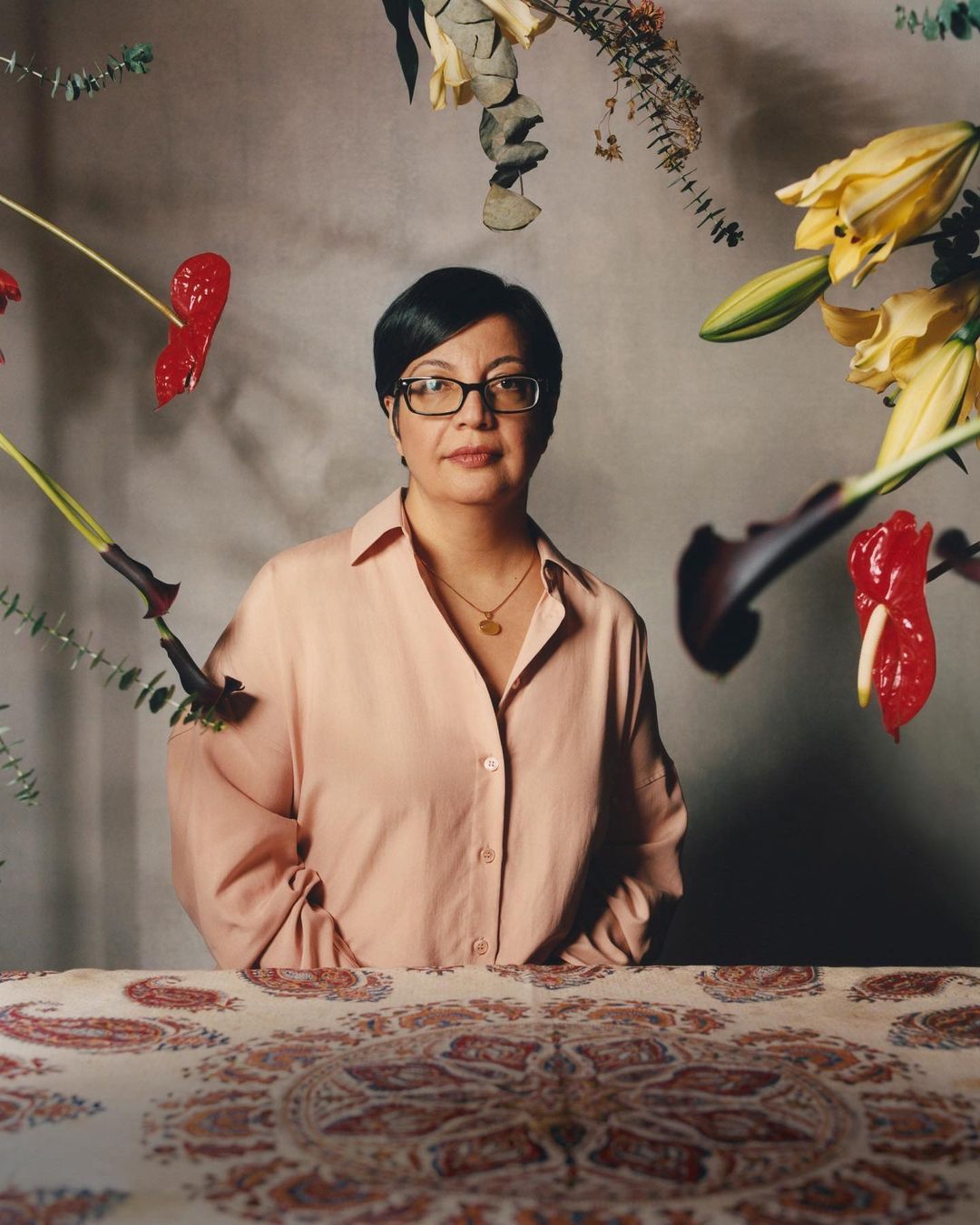 Portrait of an adult woman with short black hair and glasses. She's wearing a salmon-colored shirt and gold necklace, surrounded by lilies and anthurium flowers.