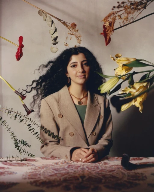 Portrait of a woman with dark, curly hair wearing a tan blazer in front of a backdrop decorated with dried flowers.