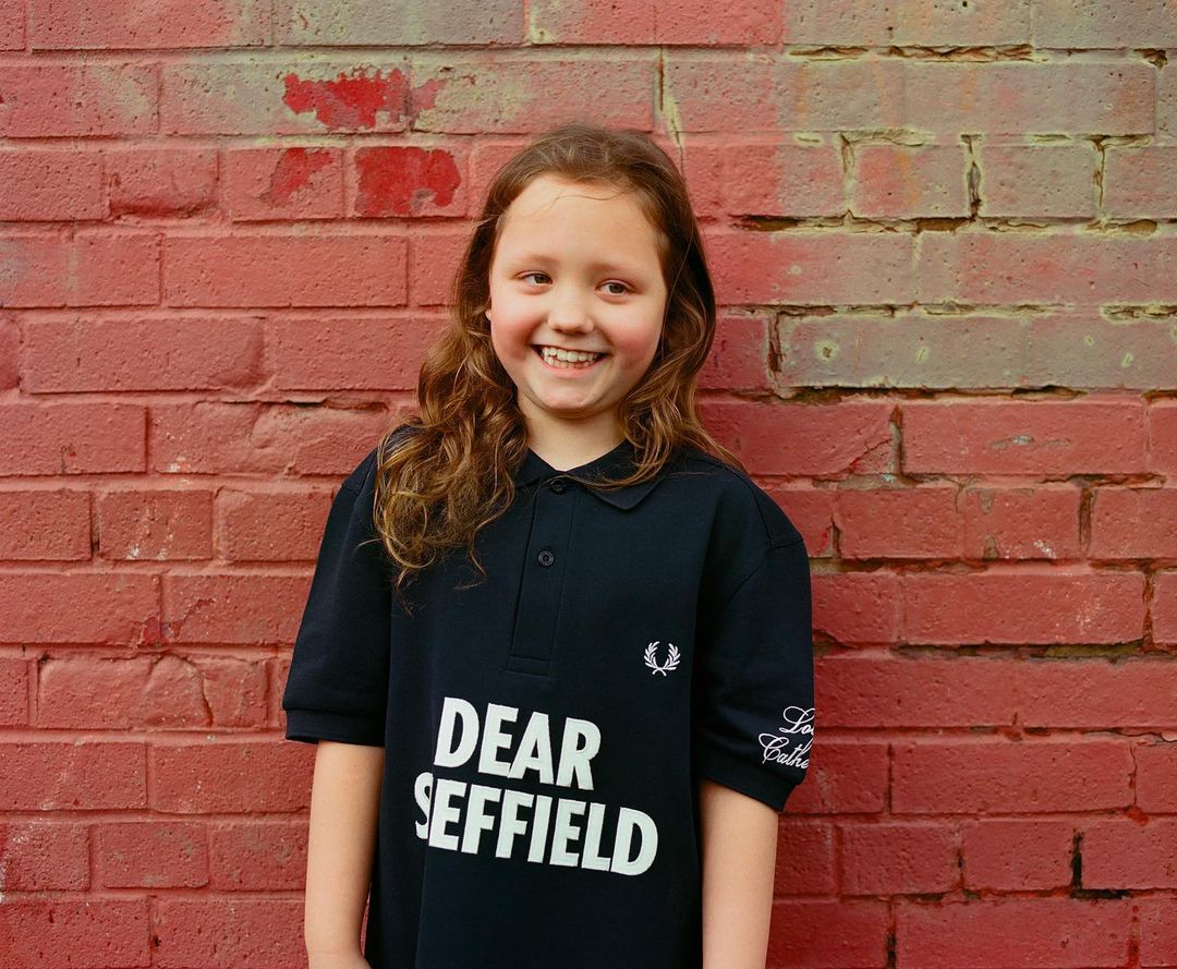 Young girl smiles in front of a red brick wall, wearing a navy polo shirt with 'Dear Sheffield' text and a Fred Perry logo.