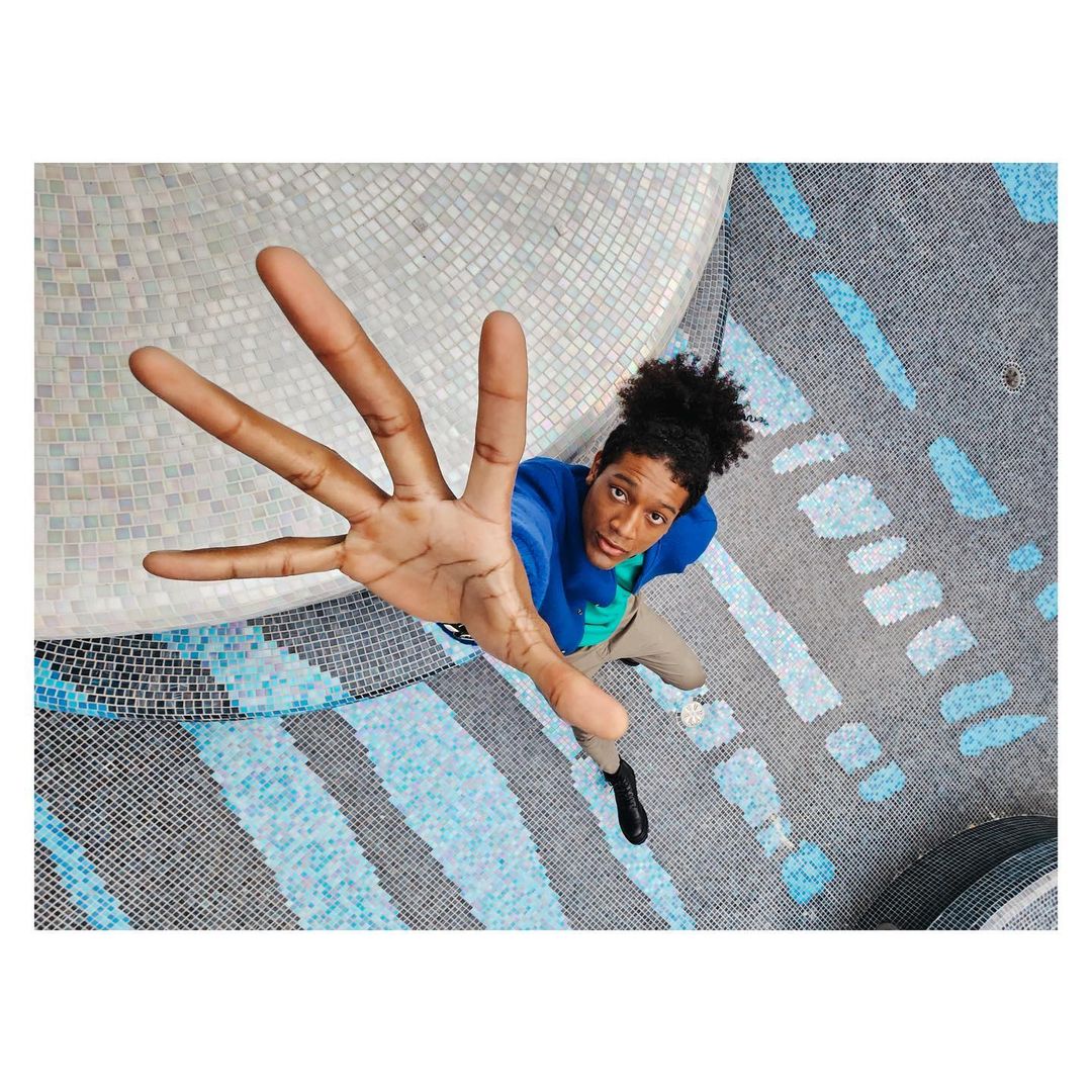 High-angle shot of a young adult with an afro wearing a blue jacket with arm outstretched, over a patterned blue and white mosaic floor.