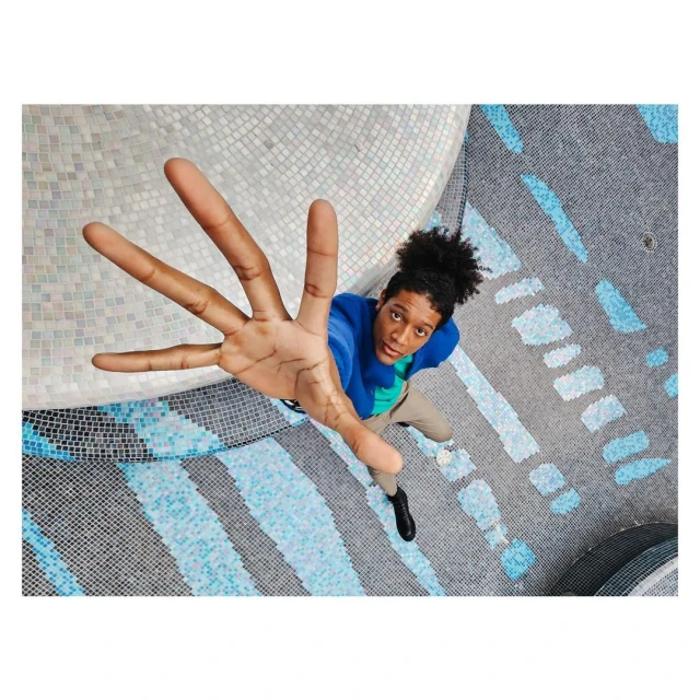High-angle shot of a young adult with an afro wearing a blue jacket with arm outstretched, over a patterned blue and white mosaic floor.
