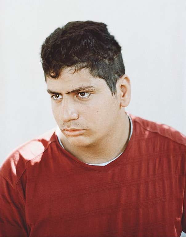 Portrait of a young adult male with dark curly hair, wearing a red t-shirt, against a simple white background in a medium shot.