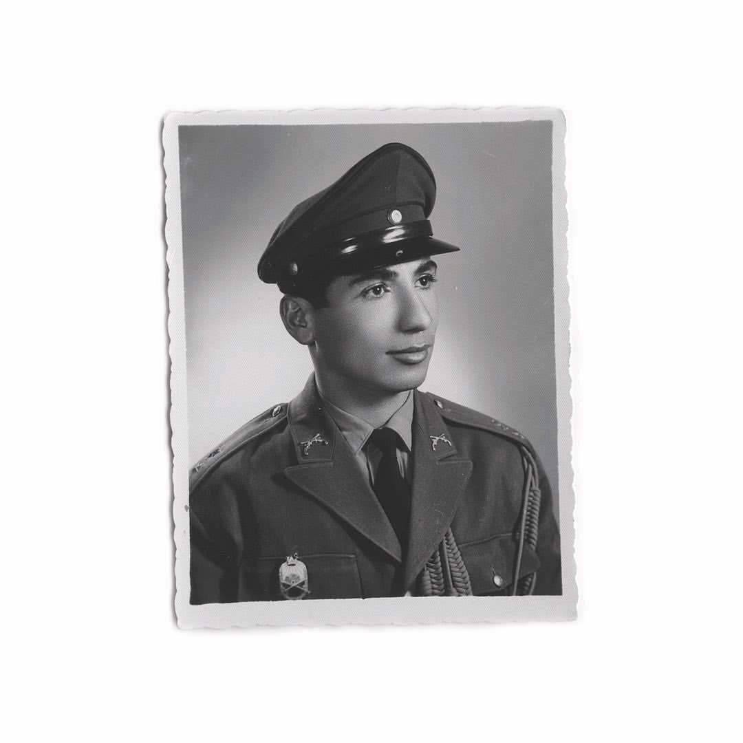 Black and white studio portrait of a young adult man in a military uniform and cap looking off to the side against a plain background.
