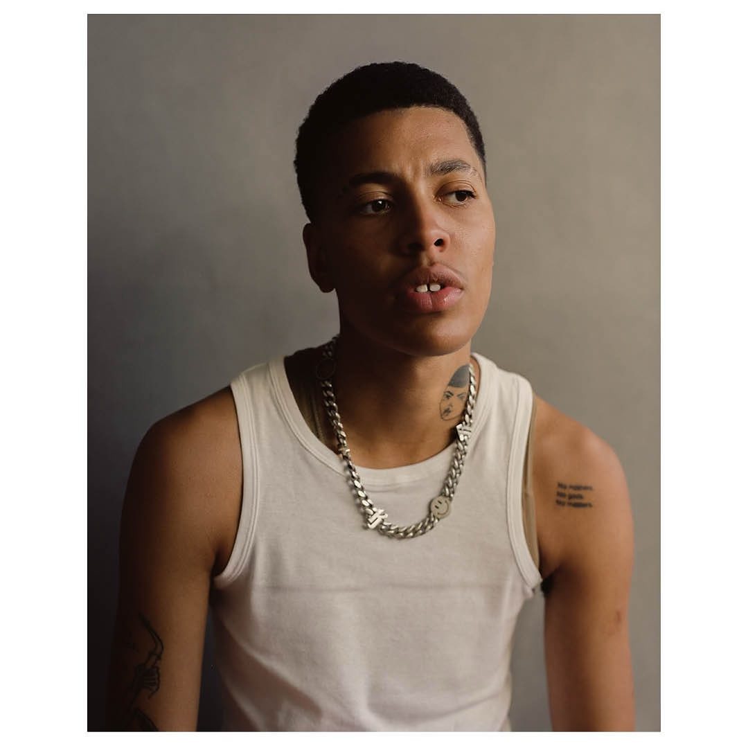 Close-up portrait of a young adult with short dark hair wearing a white tank top and a silver chain necklace against a grey backdrop.